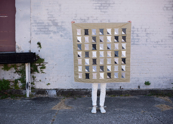 Carolyn holding Spools quilt in front of a brick wall